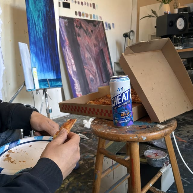An artist's studio with paint-splattered floor, canvases, a pizza box on a stool