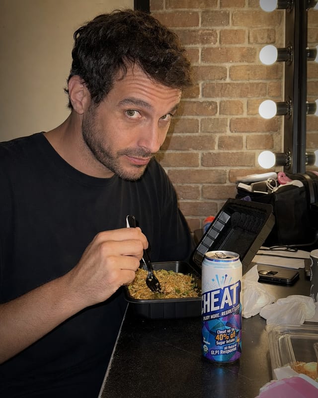 A comedian backstage in a green room, brick wall, mirror lights, takeaway box open