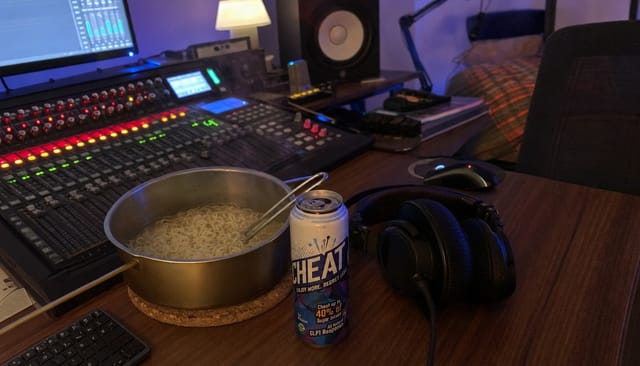 Small home studio desk at 2am, mixing console glow, headphones, late-night noodles in a pot on the desk