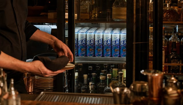 A restaurant backbar with a glass-front fridge, dim warm light, bottles, a bartender's hands