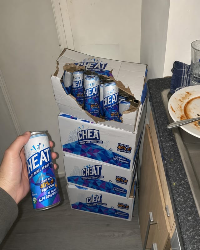 Cardboard beverage cases stacked in the corner of a small apartment kitchen, one case torn open, phone photo