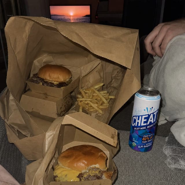 A food delivery paper bag opened on a couch, burger boxes and fries, TV glow, blanket